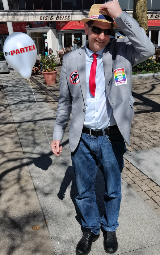Tobias Lotz im PARTEIKampfanzug mit Sonnenbrille und Hut in der Fußgängerzone in Bremerhaven. Er hält einen weißen Die PARTEI Luftballon in der rechten Hand. 