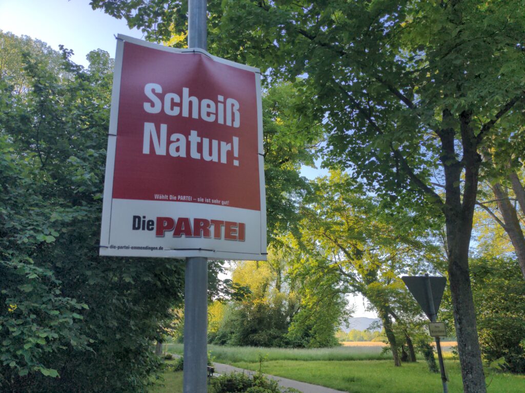 PARTEIplakat rot mit AUfdruck "Scheiß Natur!" vor grüner Wiesen und Bäumen mit Sonne.