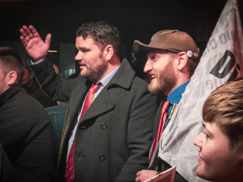 Zwei bärtige Menschen im Partei-Outfit in Profilansicht. Beide schauen in die gleiche Richtung (links). Die linke Person hebt den Arm, die Handinnenfläche ist zu sehen.