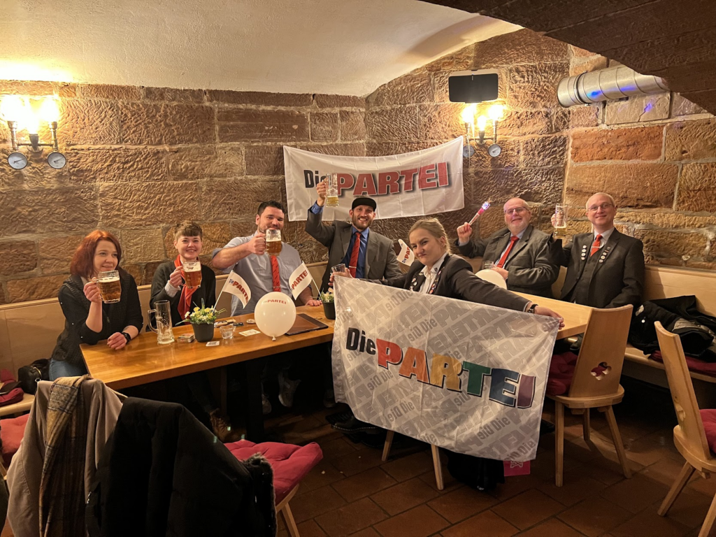 Menschen, größtenteils im seriösen Outfit eines Politiker, sitzen in einem Gewölbekeller. Biergläser und ein rosa Zauberstab werden empor und in Richtung Kamera gehalten. Im Hintergrund hängt eine Fahne der Partei Die PARTEI. vorne wird eine ebensolche Fahne von einer weiblich gelesenen Person gehalten. Der PARTEI-Schriftzug dieser Fahne ist in Regenbogenfarben gehalten.