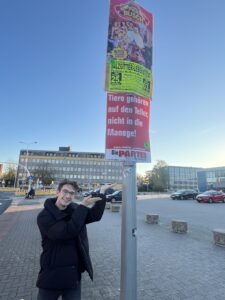 Canjoel steht vor einem Plakat auf dem steht "Tiere gehören auf den Teller, nicht in die Manege!". Darüber ist die Werbung von einem Zirkus zu sehen.